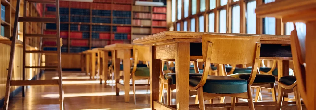 Classroom with wooden chairs and desks in rows. The room is lined with bookshelves with light coming in from floor length windows.