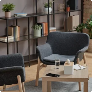 View of two dark grey lounge chairs in a lounge area with a coffee table. A grid of shelves covered in plants and books can be seen behind.