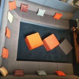 Grey U shaped sofa with small square decorative orange and grey cushions. Three square foot stools sit in the centre.