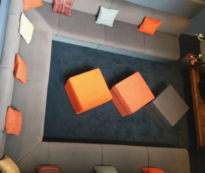 Grey U shaped sofa with small square decorative orange and grey cushions. Three square foot stools sit in the centre.