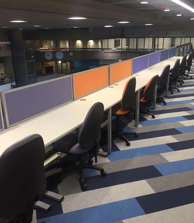 Row of desks with purple and orange boards separating them from the room. Patterned carpet flooring. 