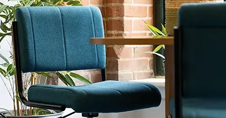 Indestructible Student Chair sat up to a desk in front of a brick wall and large plant.